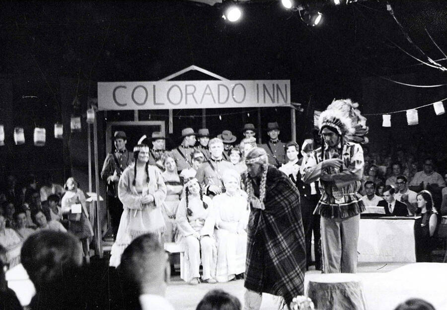 Group photo from the University of Idaho Group drama production of 'Little Mary Sunshine.' The cast can be seen dressed in costume and sitting on stage, under a sign that says 'Colorado Inn'. The audience is looking on.