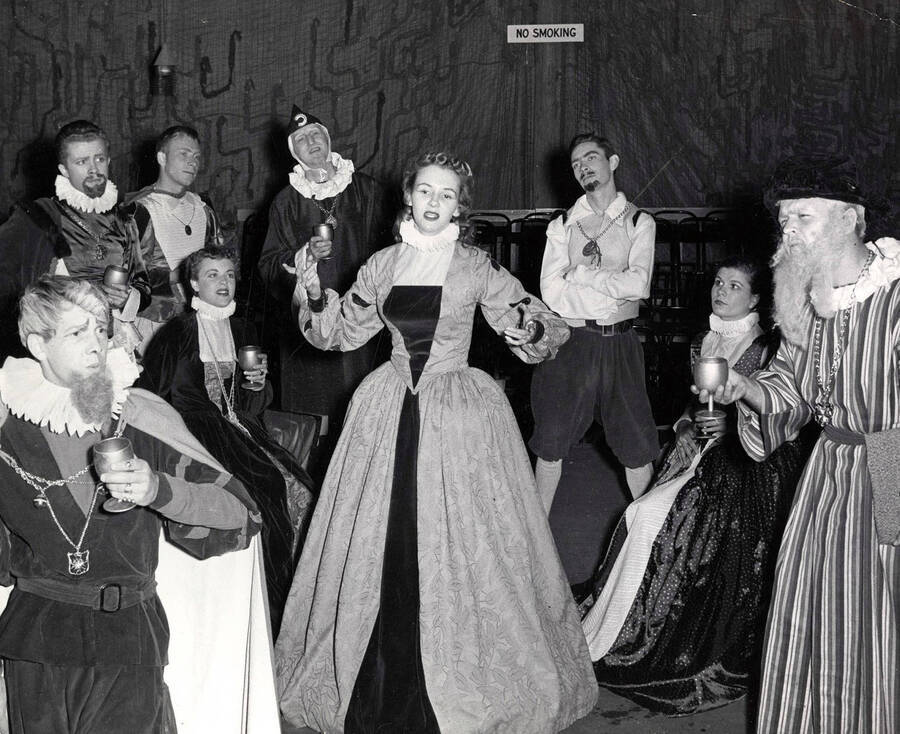 Group scene during the University of Idaho drama production of 'Taming of the Shrew.' This production was directed by Edmund Chavez. The cast members can be seen wearing costumes and holding cups.