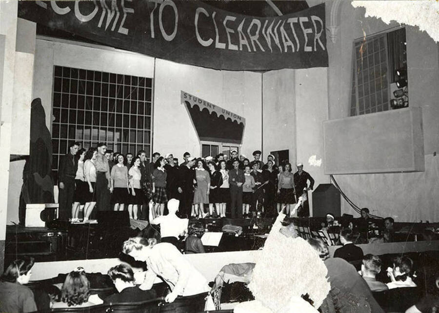 Scene from the University of Idaho drama production of 'Gee-Eyes Right.' The cast members can be seen standing on stage under a sign that reads 'Welcome to Clearwater'.