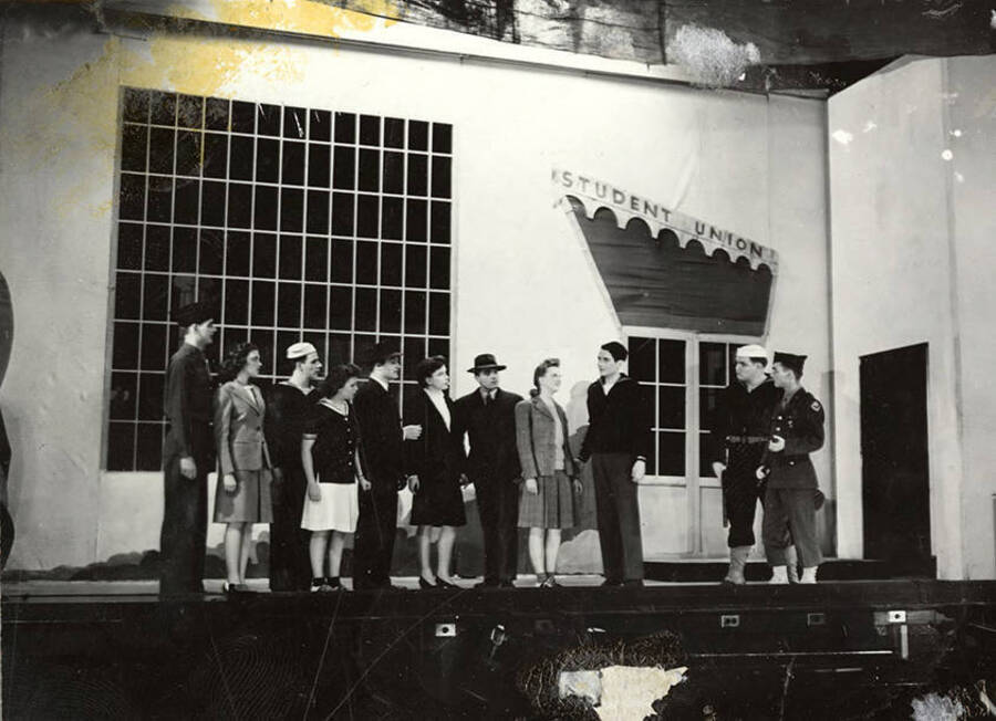 Scene from the University of Idaho drama production of 'Gee-Eyes Right.' Members of the cast can be seen standing on stage under a sign that says 'Student Union'.