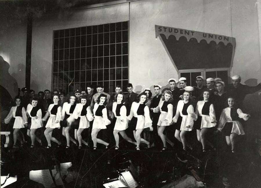 Scene from the University of Idaho drama production of 'Gee-Eyes Right.' Members of the cast can be seen standing on stage under a sign that says 'Student Union'. The women are wearing matching costumes and seem to be dancing.