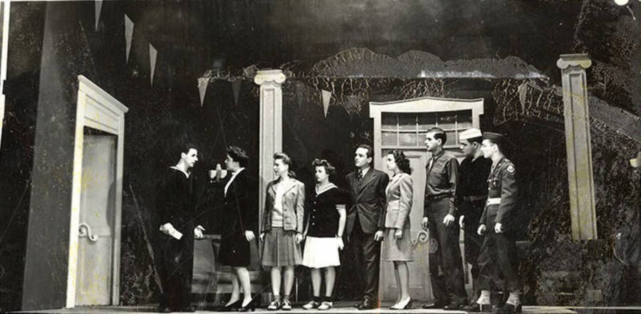 Scene from the University of Idaho drama production of 'Gee-Eyes Right.' Members of the cast can be seen standing on stage in costume, watching as a man and woman have a conversation with each other.