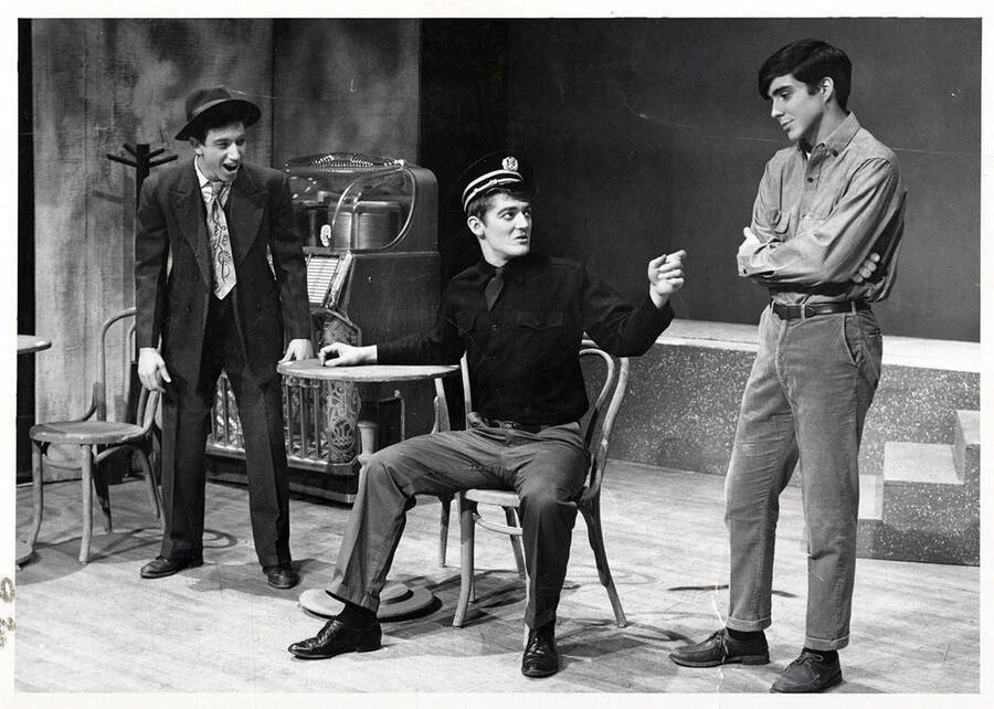 Scene from the University of Idaho drama production of 'Andorra.' This production was directed by Jean Collette. One man can be seen sitting at a table, talking with two other men.