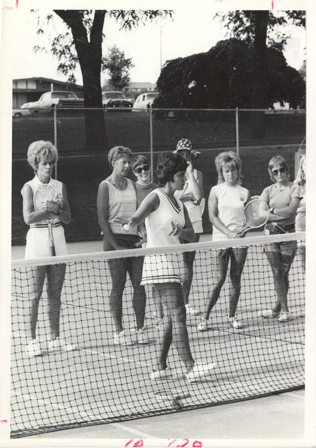 An unnamed women's Tennis instructor provides lessons while her group of players watches.