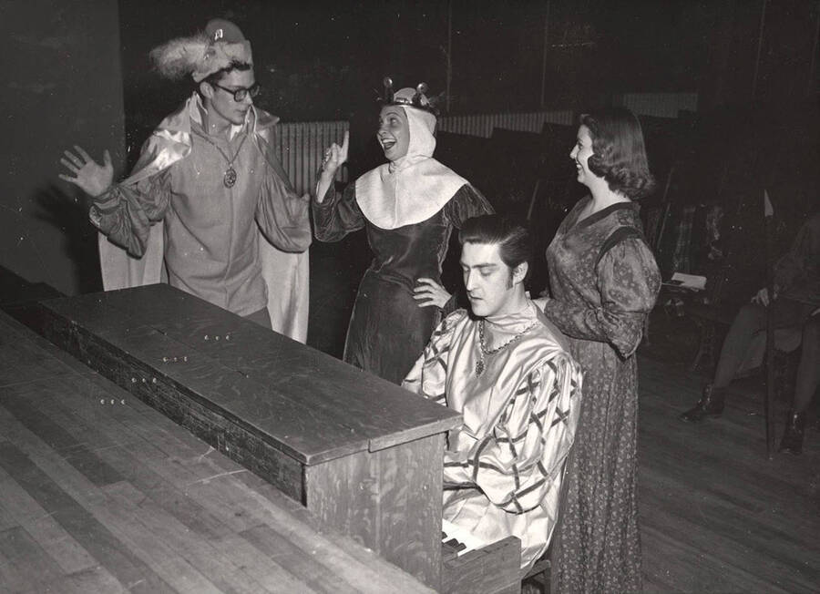 Clyde Winters as Osric, Margaret Magee as Gertrude, Charles LaFollette as Rosencrantz and Jean Bales as Lady in Waiting stand around a piano during a break from rehearsal of 'Hamlet.'