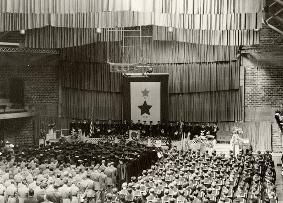 Graduates dressed in caps and gowns and military uniforms stand facing the stage during the 1945 Commencement ceremony held in the Memorial Gym.