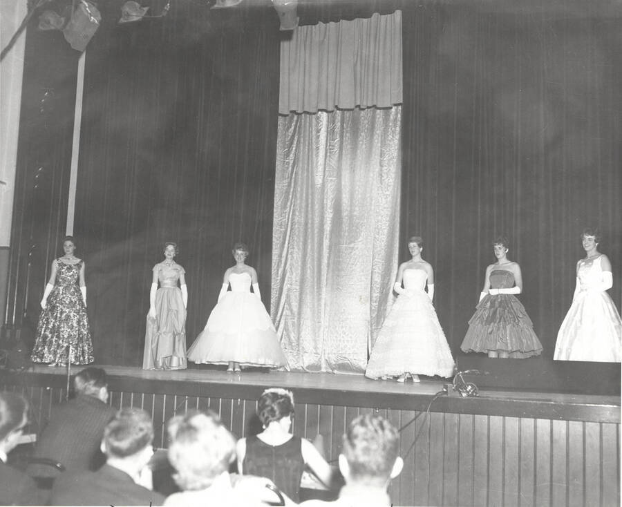 Queen contestants pose for a group photo on stage at the 37th National Convention of the Intercollegiate Knights, a national honorary service organization. Individuals identified from left to right: unknown, Donna Hilton (Brigham Young University), Queen (name unknown), Sally Newland (University of Idaho), unknown, unknown.