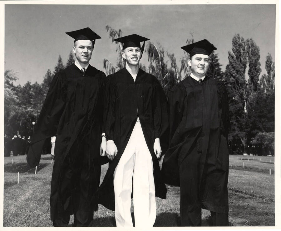 Unidentified graduates dressed in their caps and gowns stand together outdoors on a campus lawn.