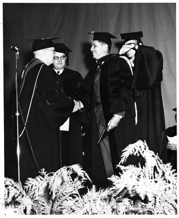 Dr. Eugene B. Chaffee receives an honorary Doctor of Laws degree from University of Idaho President Donald Theophilus during the 1964 Commencement ceremony. Dean Boyd A. Martin stands behind and Registrar Lee O'Neill places the hood on Dr. Chaffee.