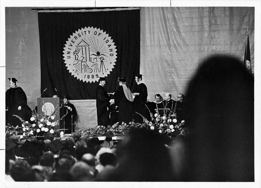 A graduate receives his degree on stage in front of a large banner of the Seal of the University of Idaho during the 1974 Commencement ceremony.
