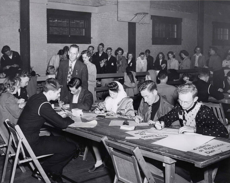 Students fill out schedule cards at tables in the foreground while students wait in line to begin the registration process in the background.