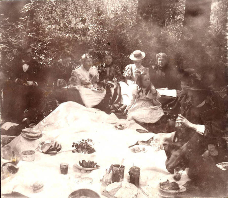 Students and their families gather for a picnic in a wooded area.