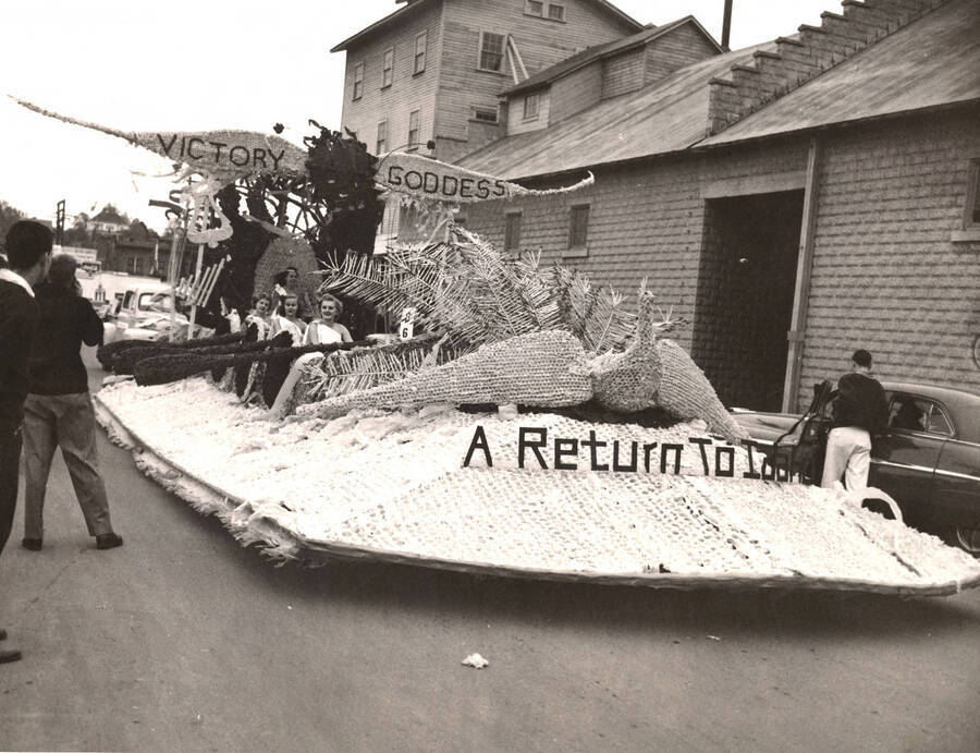 Women in togas ride on an unassociated float in the Homecoming Parade. A winged banner along the top of the float reads 'Victory Goddess' with 'A Return to Idaho' spelled out in front of a cardboard dove.