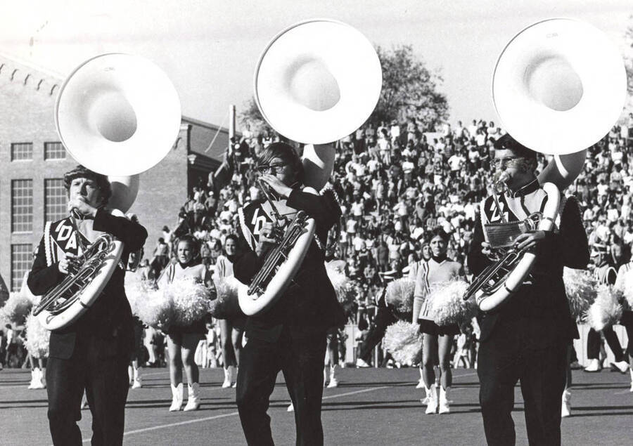 Three sousaphone players march in the half-time performance while cheerleaders encourage the team and crowd.