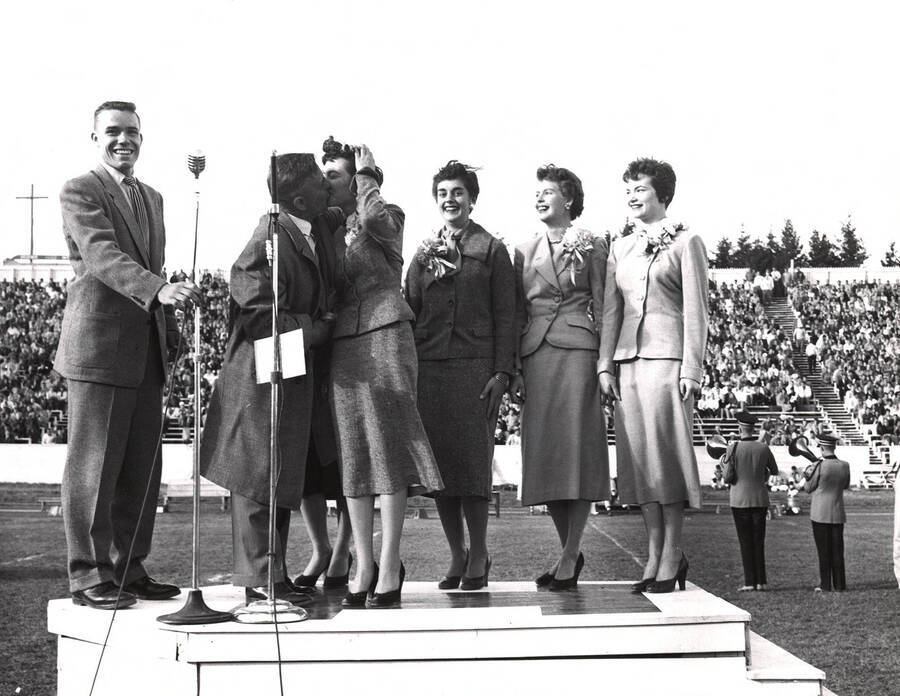 Philip Kleffner, ASUI president, holds the microphone while F.L. Bloomquist, Alumni Association President, gives Homecoming queen Clara Armstrong the traditional kiss. Attendants in second row: Sonia Henriksson, Carol Ann Zapp, Lou Ann Olson, and Freda Payne.