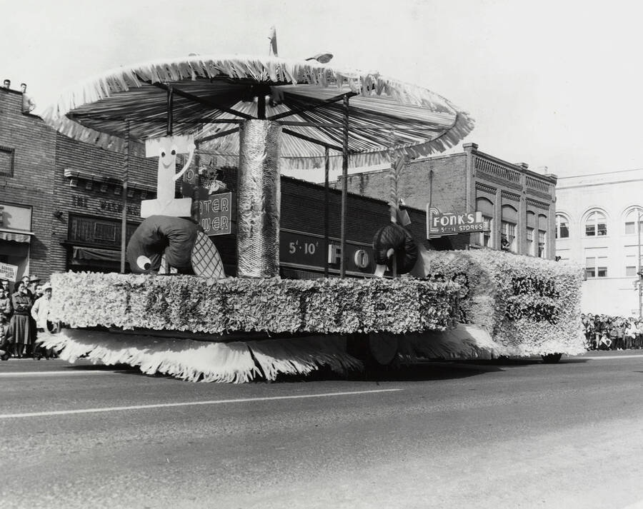 An unidentified float in front of Washington Water and Power and Fonk's during the Homecoming parade, featuring a smiling 'I' under an umbrella.