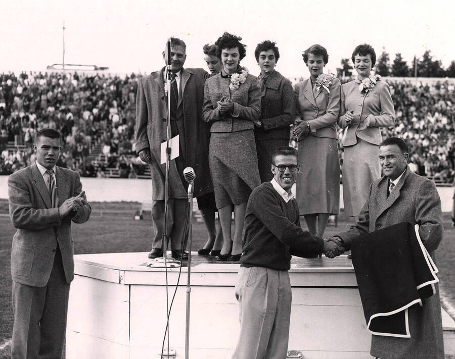 Homecoming queen Clara Armstrong and her court watch as  Earl Bullock is awarded the "I" blanket for being "Outstanding Idaho Booster." Attendants in second row: Sonia Henriksson, Carol Ann Zapp, Lou Ann Olson, and Freda Payne. Also, Philip Kleffner clapping and F.L. Bloomquist on podium.