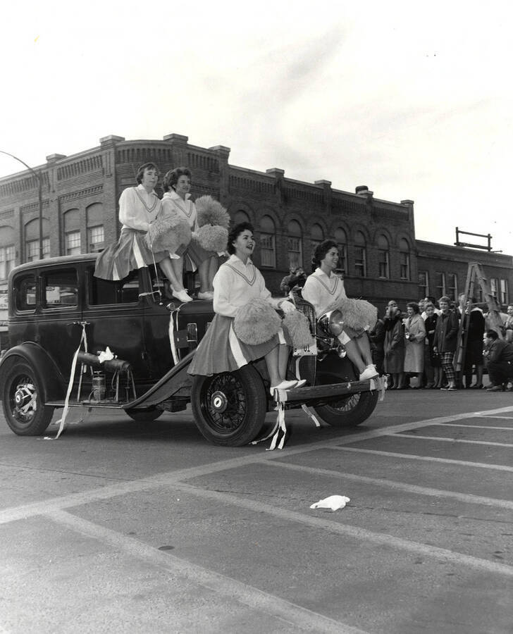 Women with pom poms ride on the front of a car during the Homecoming parade.