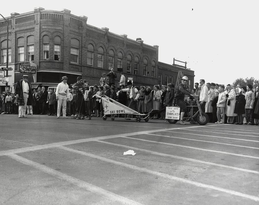 A man pulls the North South Ski Bowl sled behind his scooter with a sign saying "Tote Gote, Courtesy of Charlie's Saw Shop" during the Homecoming parade.