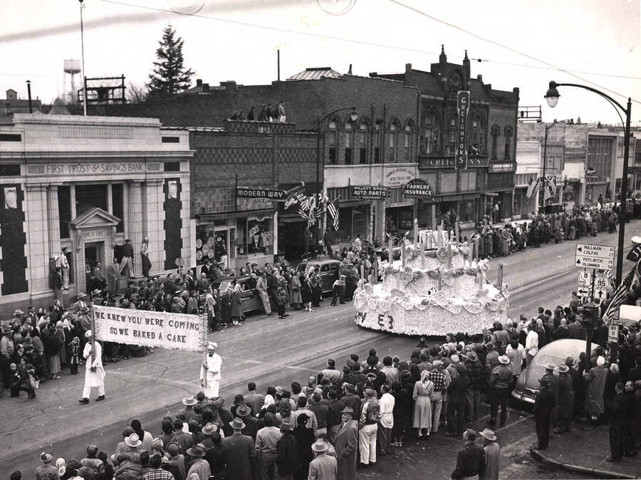 Two men carry a sign in front of Phi Gamma Delta's prize winning birthday cake float. The caption reads "We Knew You Were Coming So We Baked a Cake."