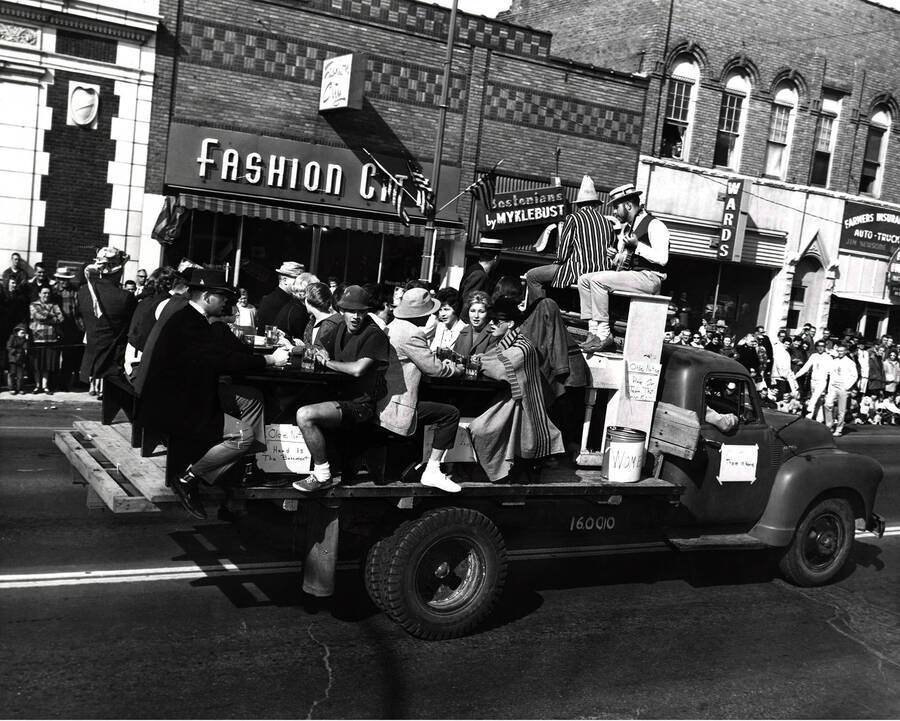A group of people sit at tables and on top of a piano as part of an unidentified float in front of Fashion City, Bostonians by Myklebust and Wards.