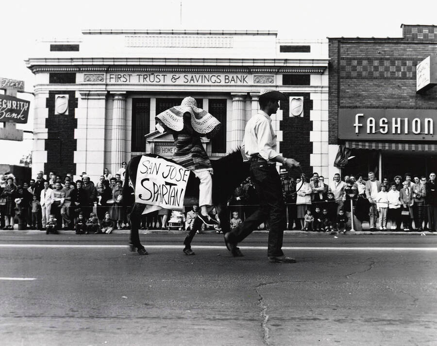 An unidentified man leads "San Jose Spartan" on a donkey during the Homecoming parade. In front of First Security Bank and Fashion City.