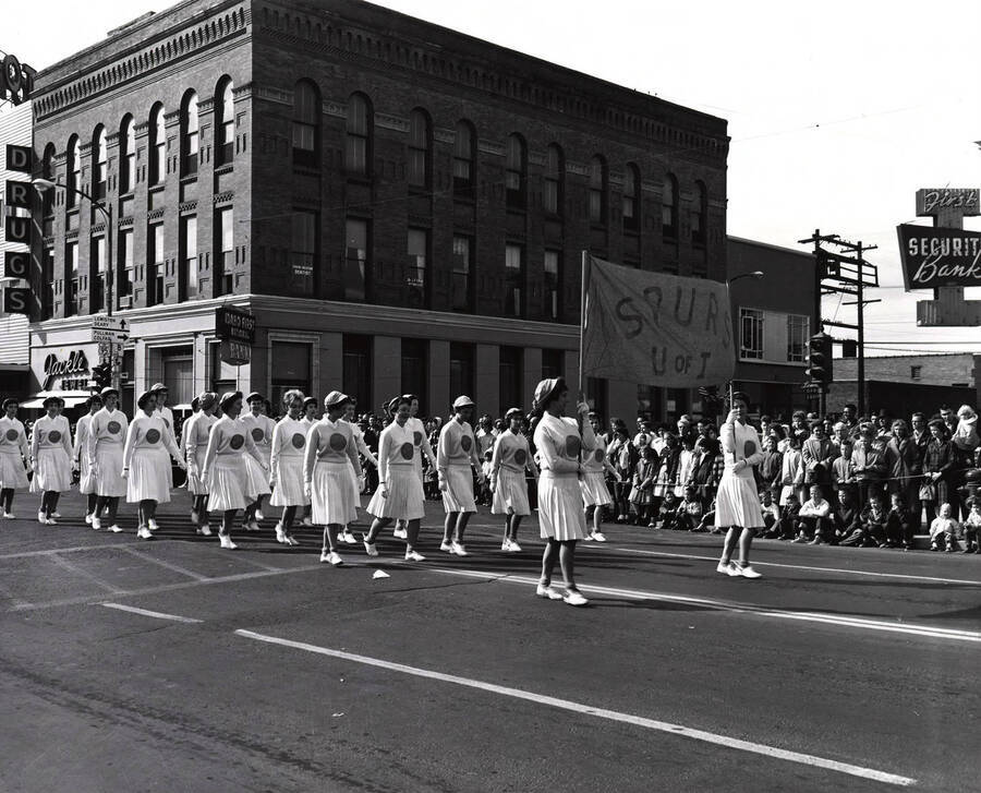 The "Spurs U of I" march in their white uniforms in the Homecoming parade in front of Jackle Jewelry and Idaho First National Bank.