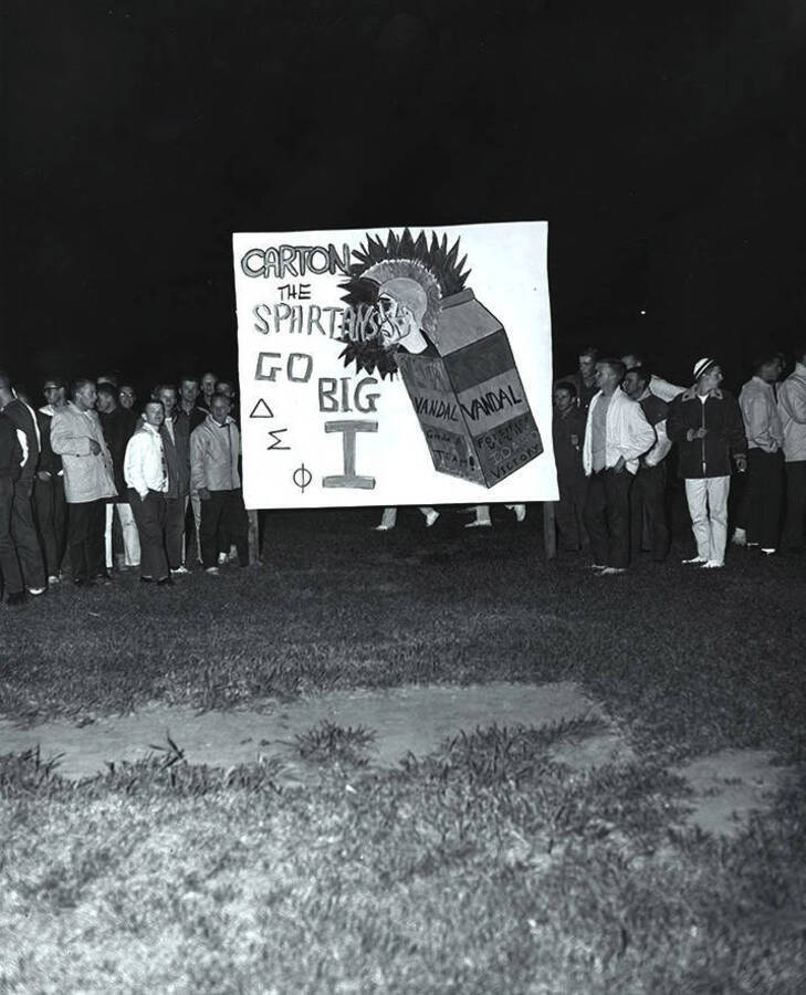 A group of students stand next to a sign that reads "Carton the Spartans Go Big I Delta Sigma Phi" at the Homecoming bonfire.