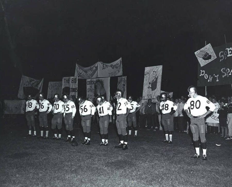 Football players stand in front of signs and spectators during the Homecoming bonfire.