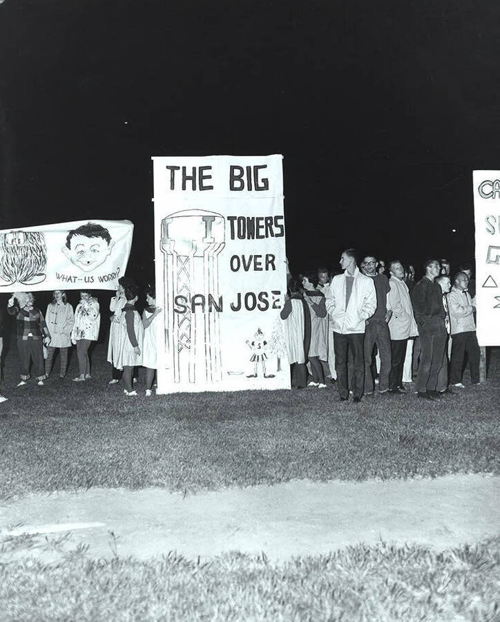 Students stand with signs reading "The Big I Towers over San Jose" and "What-Us Worry?" during the Homecoming bonfire.
