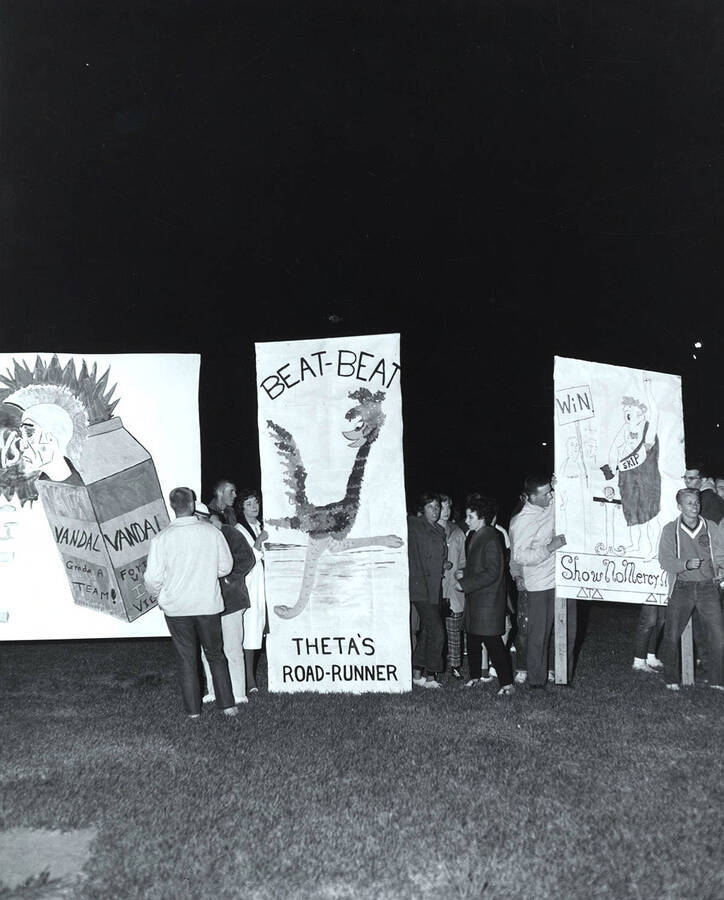 A group of students stand beside three signs reading "Beat-Beat, Theta's Road-Runner" and "Win, Show No Mercy".