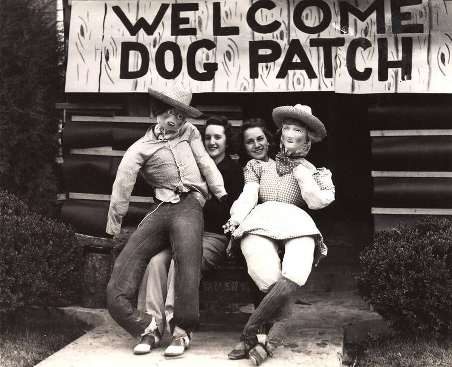 Two Delta Gamma women sit in front of the Delta Gamma Homecoming house decorations with two large dolls on their laps. The sign over the door reads "Welcome Dog Patch."