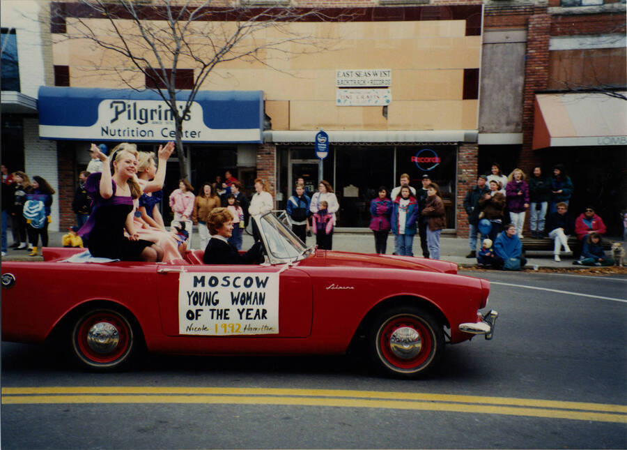 Nominees for Moscow Young Woman of the Year ride in a red car in the Homecoming parade. A sign on the side of the car reads "Moscow Young Woman of the Year Nicole Hamilton 1992."