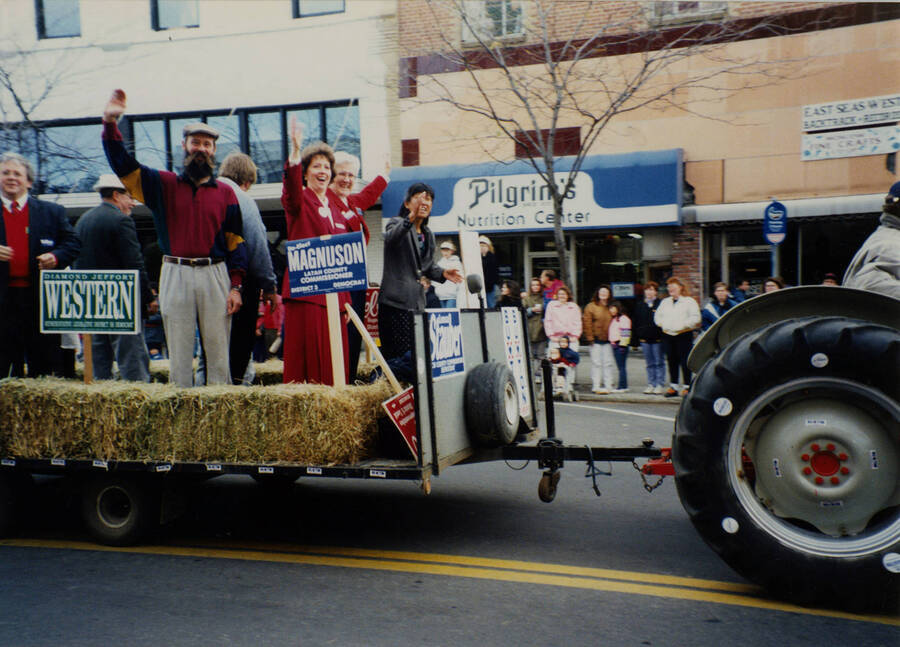 Men and women wave at spectators from behind hay bales and signs for local and county government seats as part of a float in the Homecoming parade.