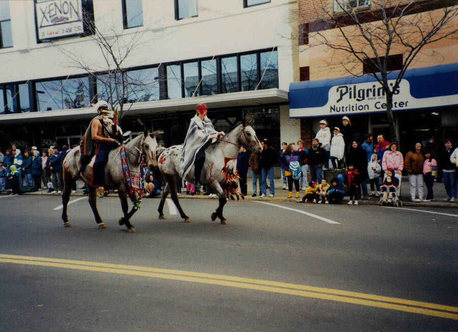 Two individuals dressed in Native American costume ride Appaloosa horses in the Homecoming parade.