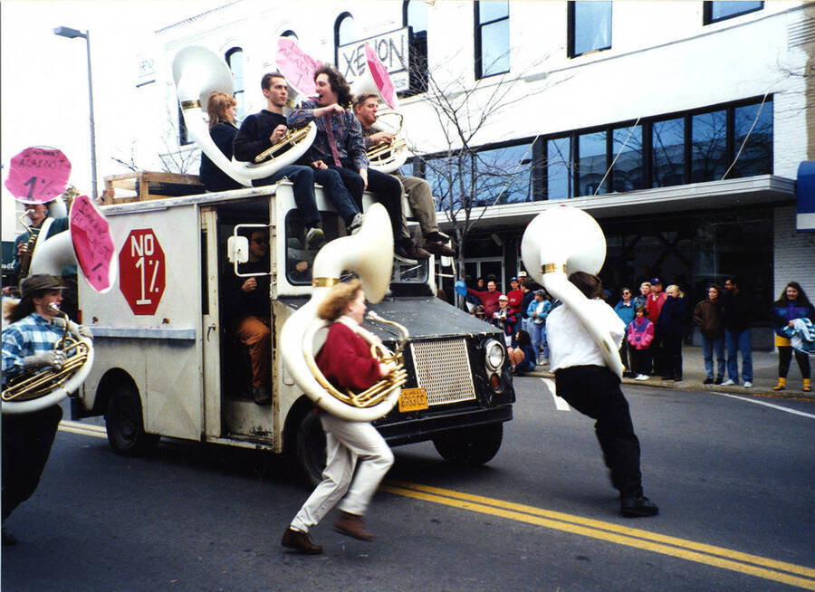 The tuba line of the marching band drive, ride, and run around a retired mail vehicle while carrying their sousaphones as part of the homecoming parade.