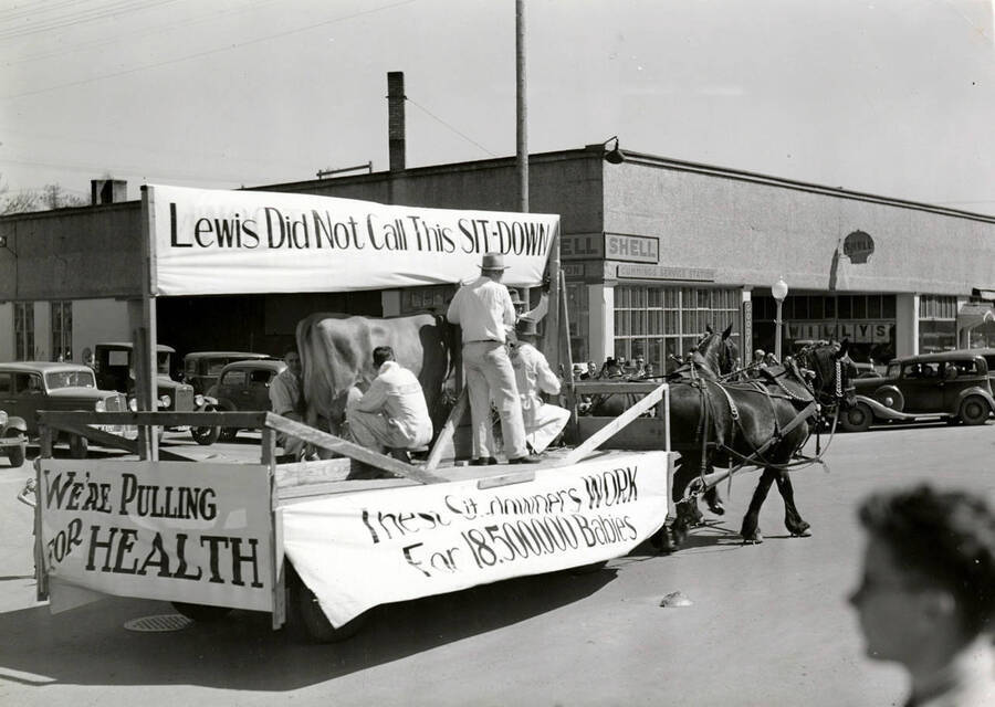 Horses pull a float in front of the Cummings Service Station, showcasing men milking a cow and signs that read, 'Lewis Did Not Call This SIT-DOWN,' 'We're pulling for Health' and 'These sit-downers WORK for 18.500.000 Babies' during Idaho's Little International Agriculture Show.