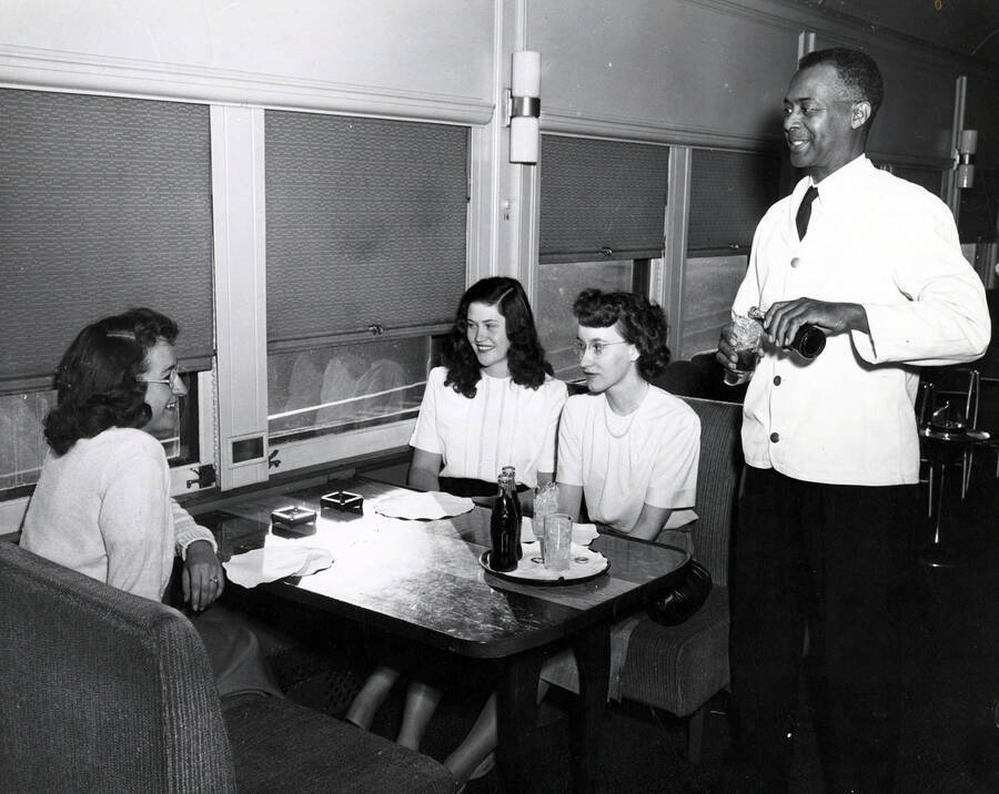 A waiter pours Coca-Cola for three students who talk while seated in a club car of the Student Special train.