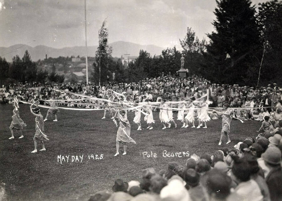 Students dressed in light wear carry poles overhead during Idaho's Campus Day celebrations. Caption reads: "May Day 1925 Pole Bearers."