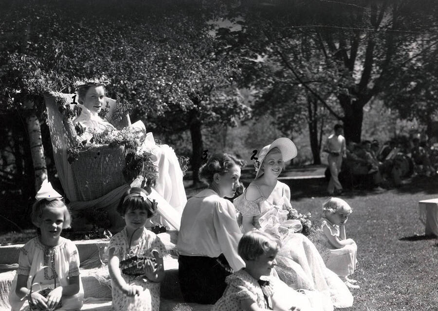 A side-angle of Idaho May Queen, Margaret Brodrecht, as she sits on a throne surrounded by her court for the Campus Day celebrations. Students pictured: 1) Margaret Brodrecht (May Queen), 2) Jane Baker (Maid of Honor), 3) Donna-May South (page), 4) Ann DuSault (flower girl).
