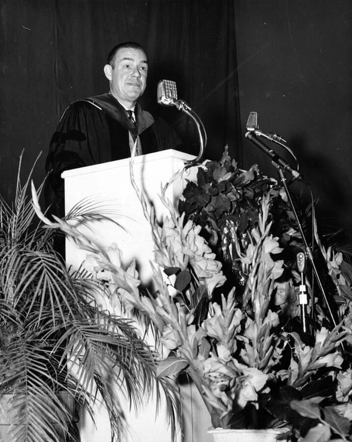 Governor Robert E. Smylie speaks at Idaho's commencement on a podium surrounded by flowers.