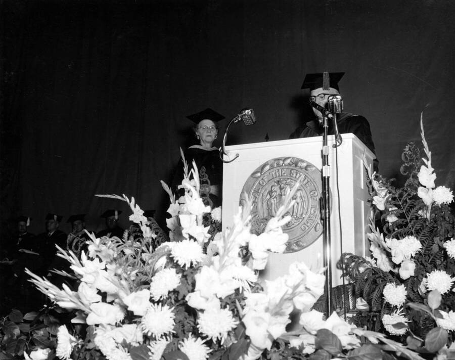 University of Idaho President Donald R. Theophilus reads the Citation of Merit from a podium with flowers in front after Dean of Women Louise Carter retired.