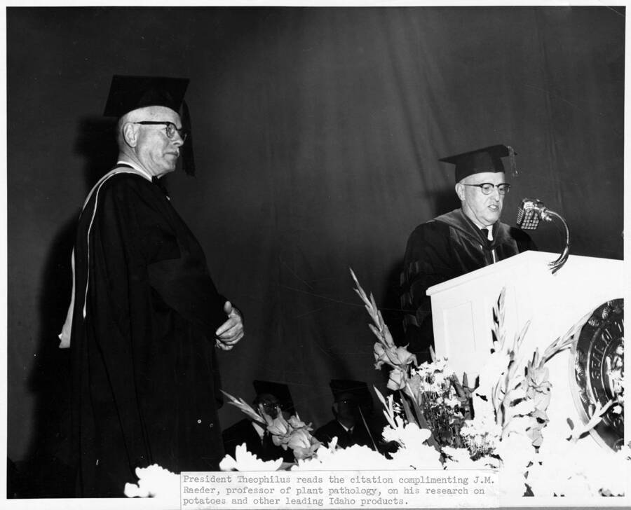 University of Idaho President Donald R. Theophilus stands at the podium, congratulating Raeder during University of Idaho's commencement. Caption reads: 'President Theophilus reads the citation complimenting J.M. Raeder, professor of plant pathology, on his research on potatoes and other leading Idaho Products.'