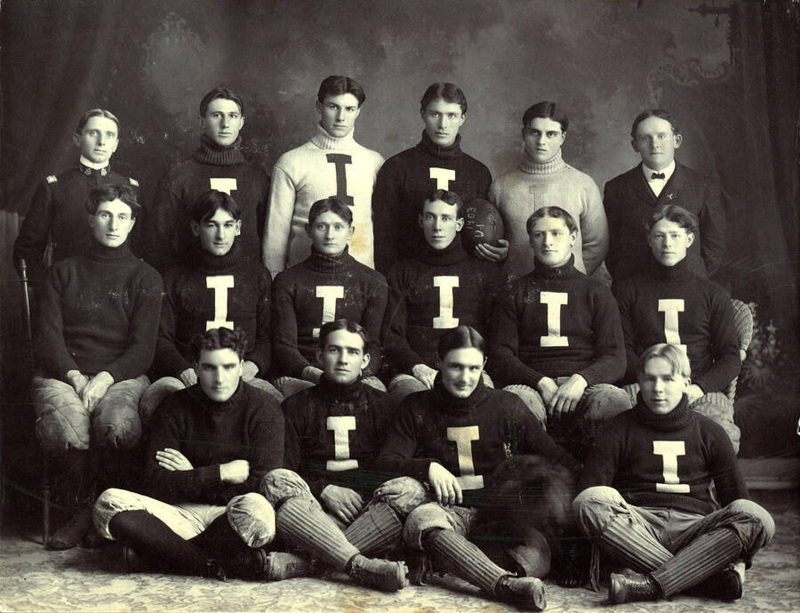 A group picture of the University of Idaho football team in turtleneck sweaters with a large 'I' decal. Pictured are: Back) Captain Chrisman, Clyde Oaks, Arthur Rogers, Harry C. Smith, Arthur Strong, Coach John C. Griffith; Center) Louis Fogel, John H. Miller, John Middleton, William Snow, Gus Larson, Charles Smith; Front) Elmer Armstrong, Bill Thomas, Gray, Tilley