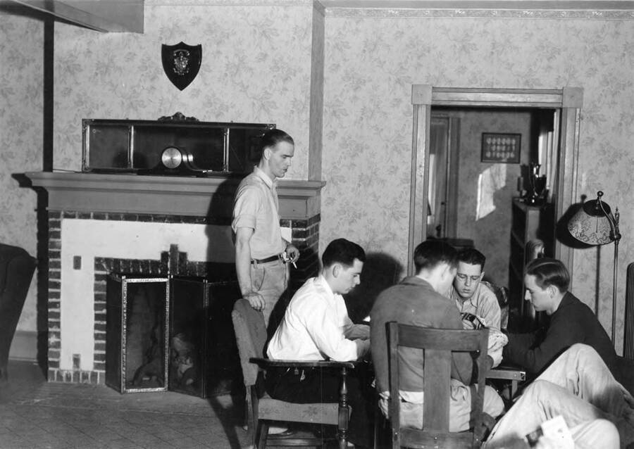 Students are sitting around a table, playing cards in the living room of the Lambda Chi Alpha house.