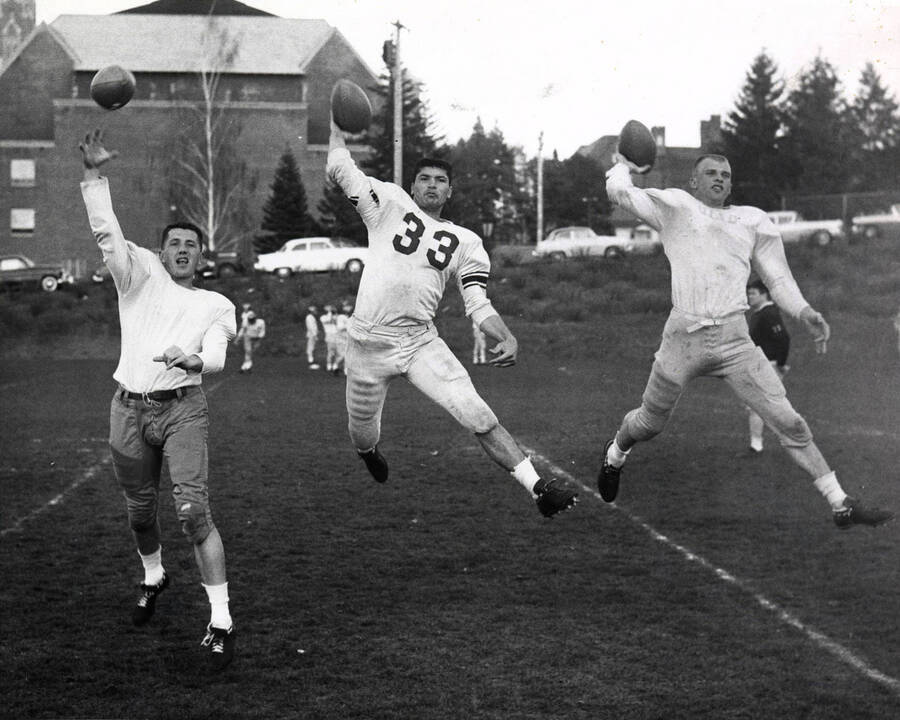 Football players identified from left to right: Sil Vial, Joe Espinoze, and Gary Farnworth jumping in the air to throw footballs at the University of Idaho.