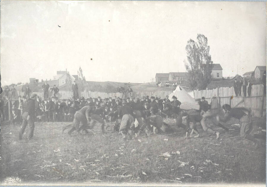 An unidentified runner is gang-tackled on what appears to be a fall afternoon of football. Fans can be seen close-by, lining the field and sitting on a perimeter fence.