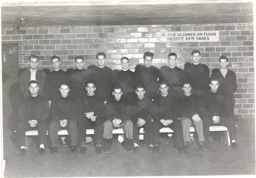 The freshman football team poses wearing all black in a locker room at Memorial Gymnasium. Players pictured: Back) Coach Otto Anderson, Rich, Rettig, Gamble, Erickson, Bowker, Gannon, Palmgren, Manager Louis Paskin; Front) Wetherall, Sanner, Betts, Pierce, Santer, Ahlskog, Fehr, Croy.