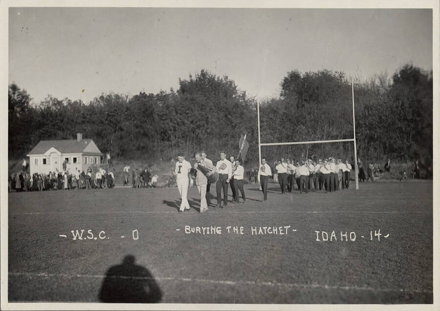 Idaho and Washington State students participate in the pregame 'Bury the Hatchet' ceremony, signifying friendship until kickoff. Caption reads 'W.S.C.-0 ~Burying the Hatchet~  Idaho-14.'