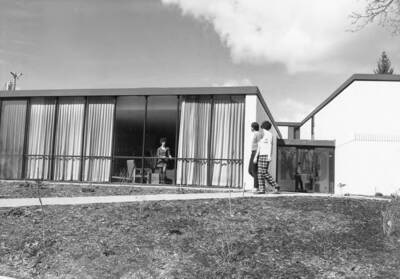 Students walk along a path outside the Kappa Alpha Theta house. Another student looks out through the large windows of a sitting room.